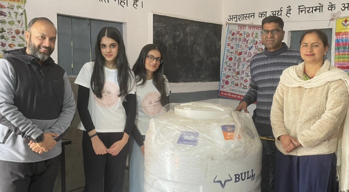 Healing Hearts Foundation team members and school staff standing beside the donated white water tank inside a classroom at Bangloo Ki Kandi Government School, with a Hindi alphabet chart on the wall behind them.