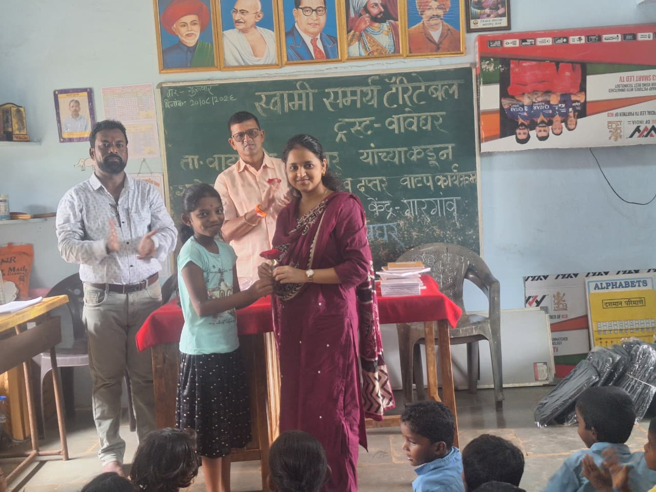 A schoolgirl presenting a rose to a woman in a classroom while students applaud, with a chalkboard and framed portraits behind them.