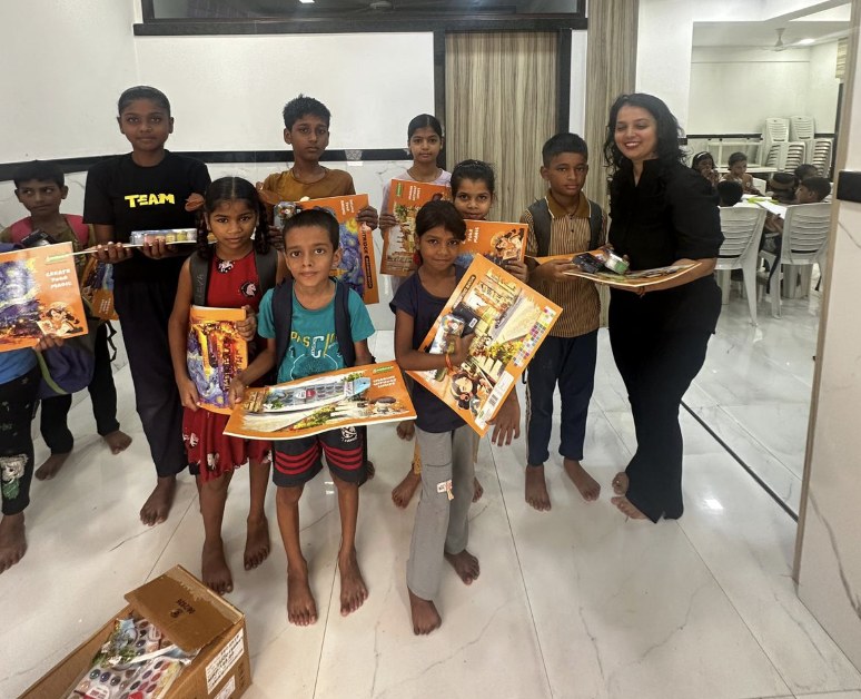 Children and an adult standing together indoors, holding up the donated drawing books, watercolour sets and geometry boxes.