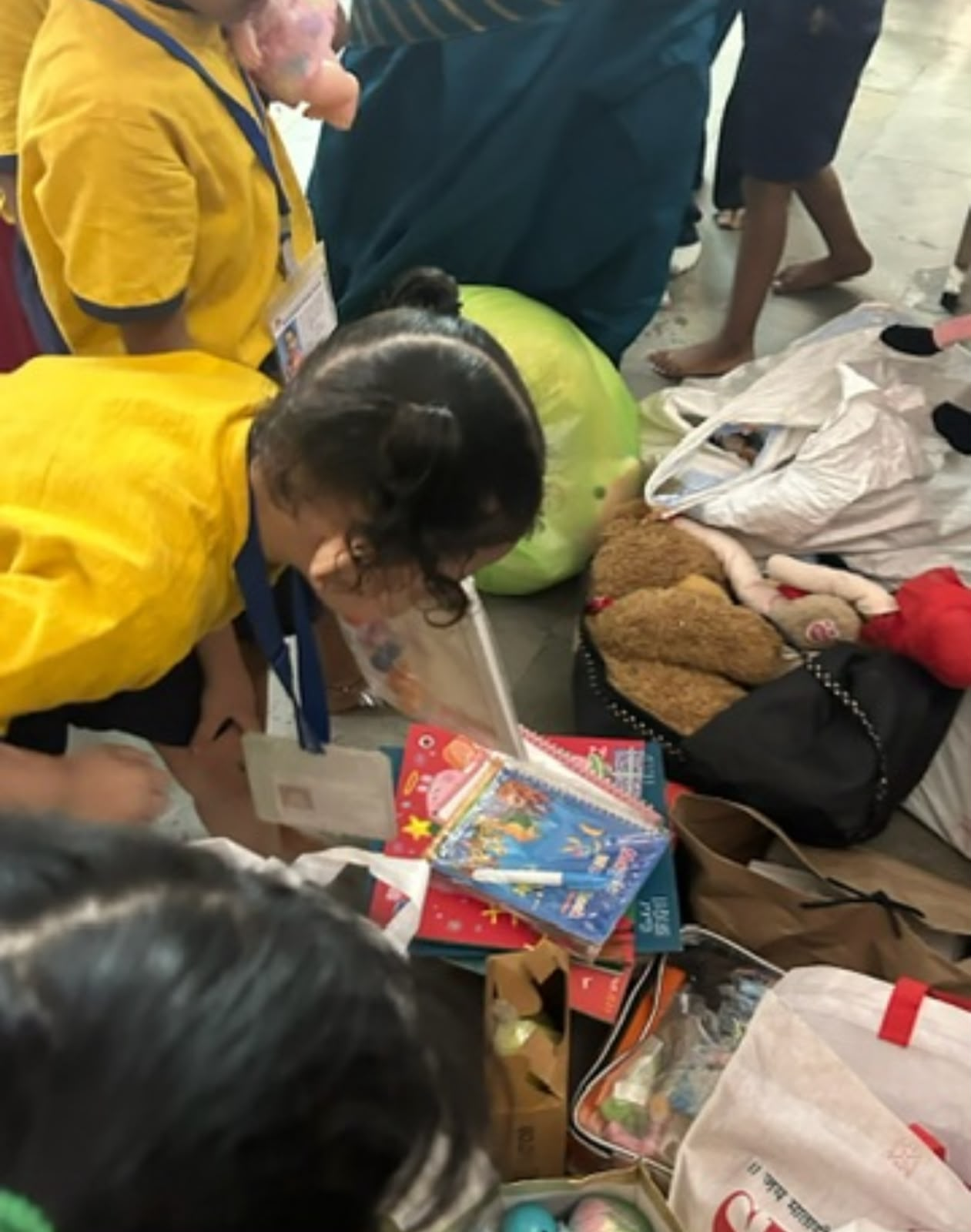 Children gathered around the donated books and toys on the floor, a girl in a yellow uniform leaning over a stack of colourful notebooks.