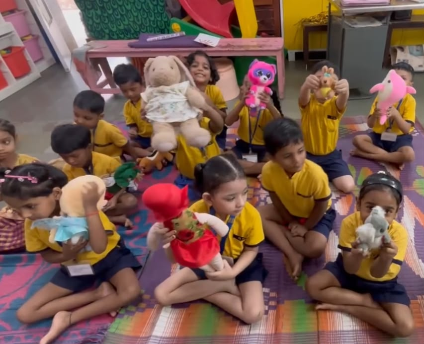 Schoolchildren sitting cross-legged on a woven mat in a classroom, smiling and holding up the stuffed toys donated by Healing Hearts Foundation.