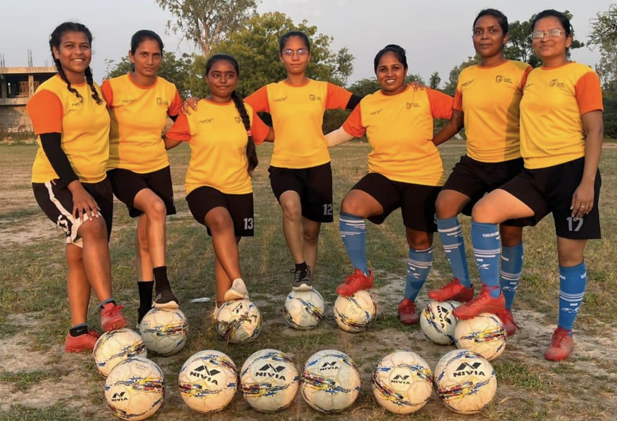 A girls' football team in yellow and orange kits standing in a line on a sports field, feet resting on soccer balls.