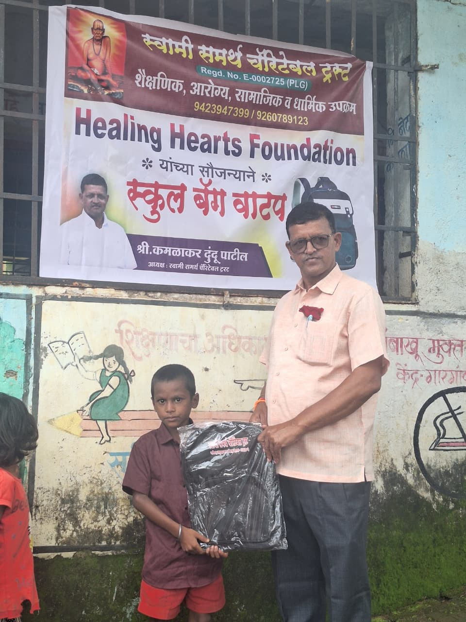 A member of the trust handing a packaged school bag to a young boy standing outside the school, with the Healing Hearts Foundation banner behind them.