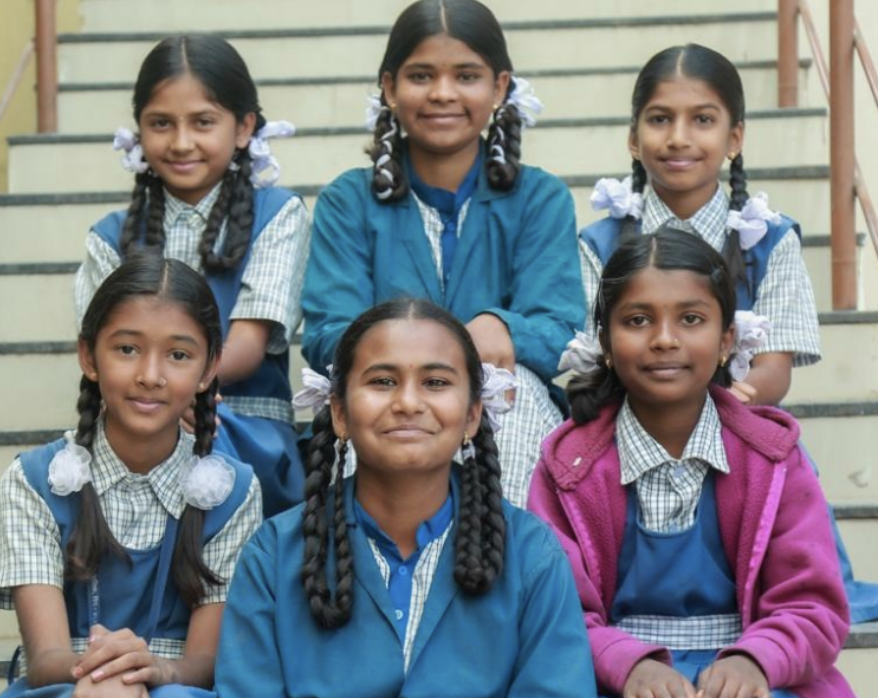 Six schoolgirls in blue uniforms with braided hair sitting together on steps, smiling at the camera.
