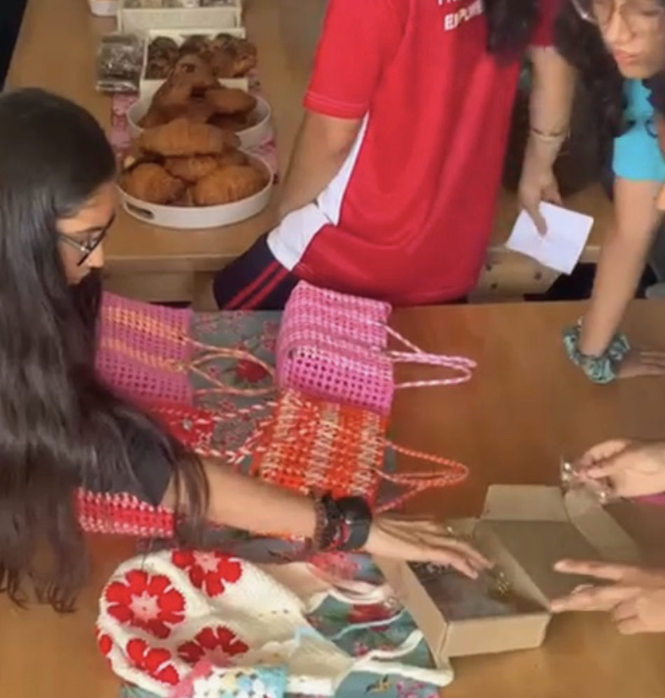 Volunteers and visitors gathered around the bake sale table, with woven pink tote bags, crocheted bags, croissants and packaged brownies on wooden tables.