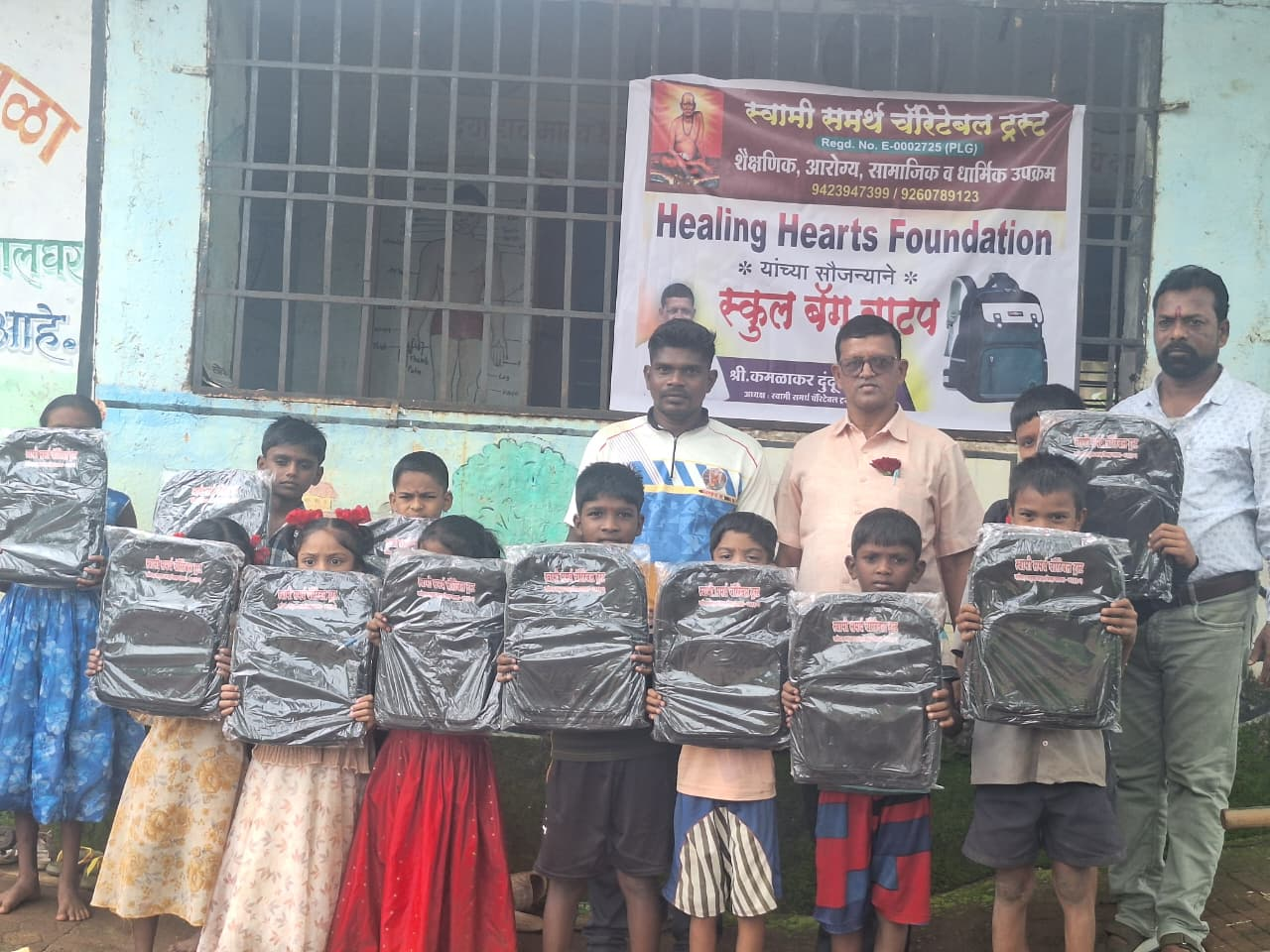 Children and members of Swami Samarth Charitable Trust and Healing Hearts Foundation standing outside a school, holding up the donated school bags beneath a school bag distribution banner.