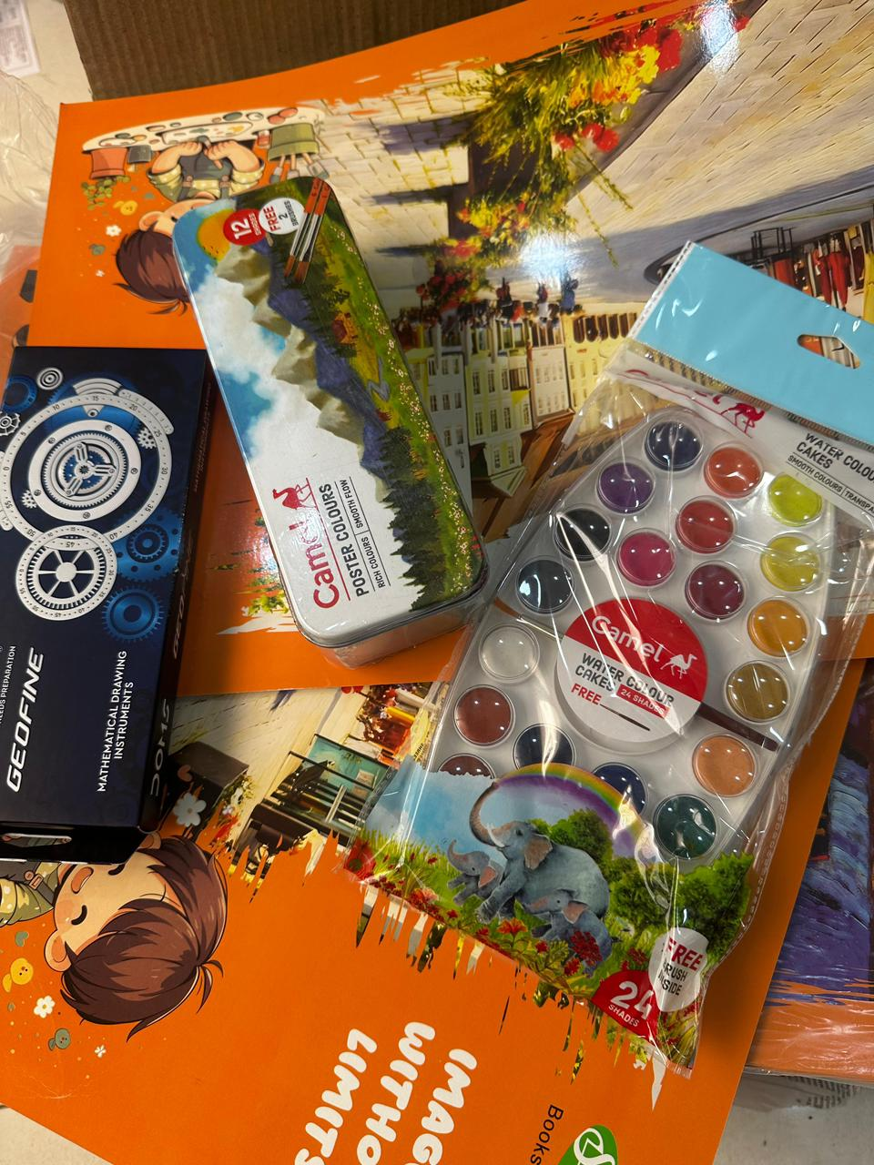Flat-lay of the donated educational supplies: an orange drawing pad, a box of DOMS Geofine mathematical instruments, a tin of Camel poster colours and a palette of watercolour cakes.