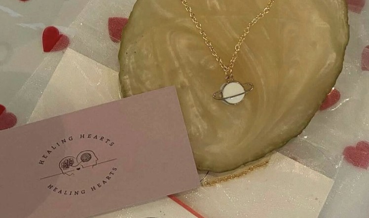 Gold chain necklace with a Saturn pendant resting on a gold resin dish beside a Healing Hearts card.