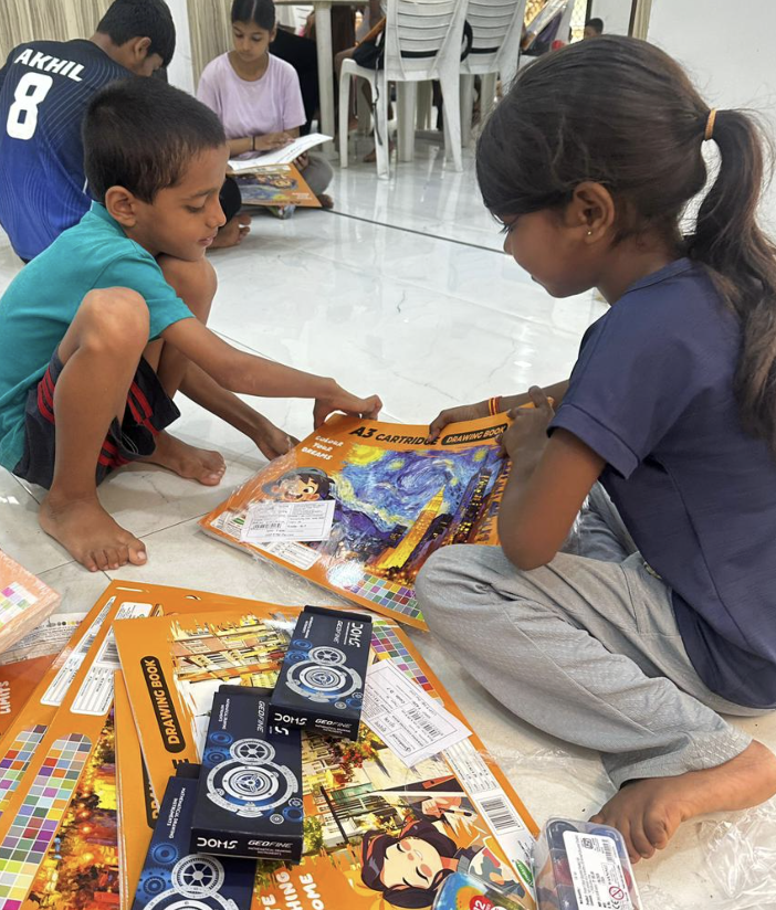 Two children sitting on a white tiled floor examining a donated A3 cartridge drawing book, with stacked orange drawing books, geometry boxes and a watercolour set in the foreground.