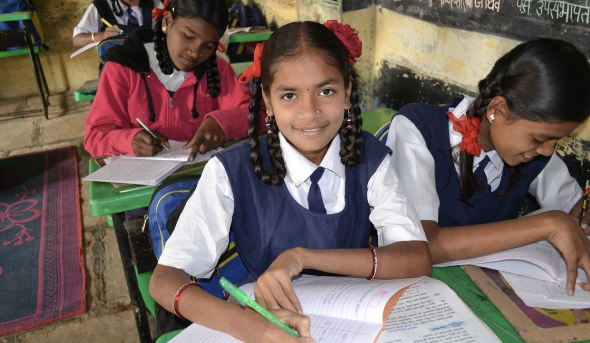 Three schoolgirls in uniform seated at green classroom desks writing in notebooks, one smiling at the camera.